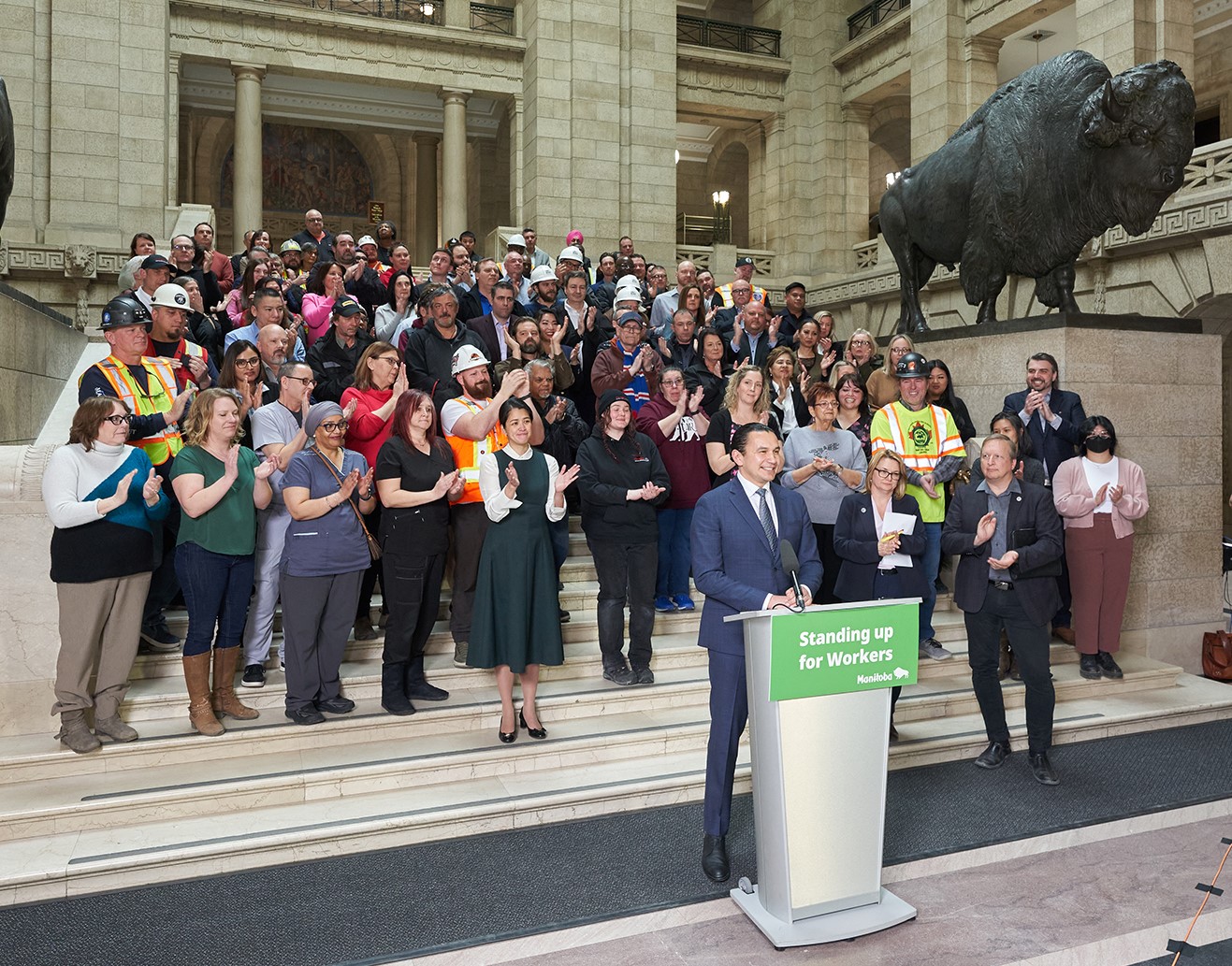 Picture of Manitoba Premier, Wab Kinew, at microphone on steps of legislature with a crowd behind him.