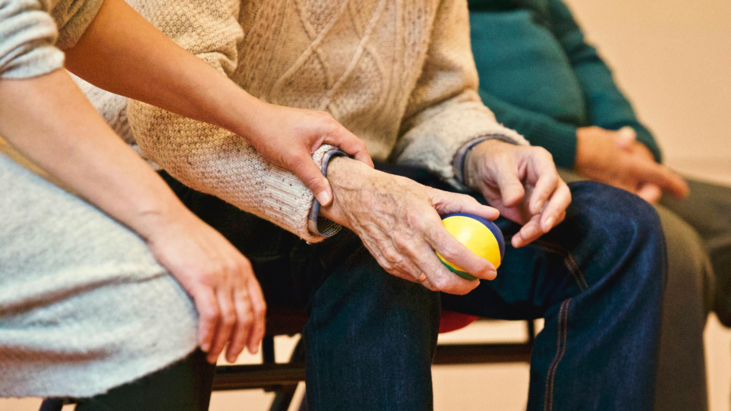 Image is a close up on the torso and hands of a young woman and an elderly man. The elderly man holds a ball in one hand. The young woman has a gentle grip on the elderly man's arm.